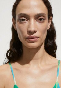 Ritratto in primo piano di una persona con capelli mossi e scuri, pelle liscia e trucco neutro, che indossa un indumento a fantasia verde e blu con spalline sottili.
