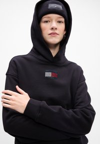 Czarna bluza z kapturem i ściągaczami, zdobiona kolorowym wzorem flagi z dżetów. Noszona z dopasowaną czarną czapką beanie.