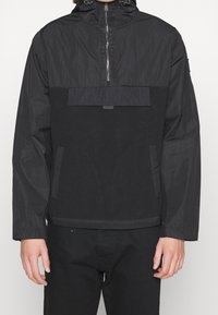 Chaqueta de pullover negra con cierre de media cremallera, secciones de tejido texturizado y un bolsillo frontal con tapa. Incluye capucha y puños ajustables.