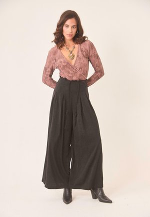 Mujer con blusa marrón atada, pantalones negros de pierna ancha con cinturón, botas negras de tacón y un collar grande de flores, posando frente a un fondo liso.