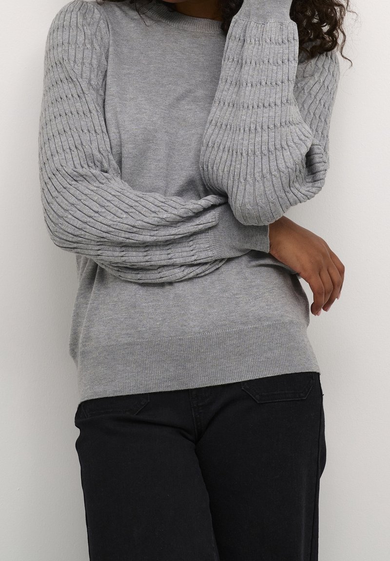 Person wearing a gray textured long-sleeve sweater with puffed sleeves and black pants, posed with arms crossed against a white wall.