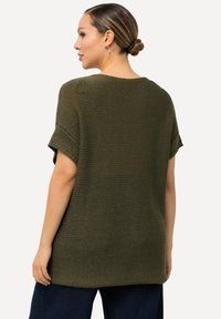 Olivgrünes gestricktes Oberteil mit kurzen umgeschlagenen Ärmeln, strukturiertem Muster und lockerer Passform, kombiniert mit dunkelblauen weit geschnittenen Hosen.