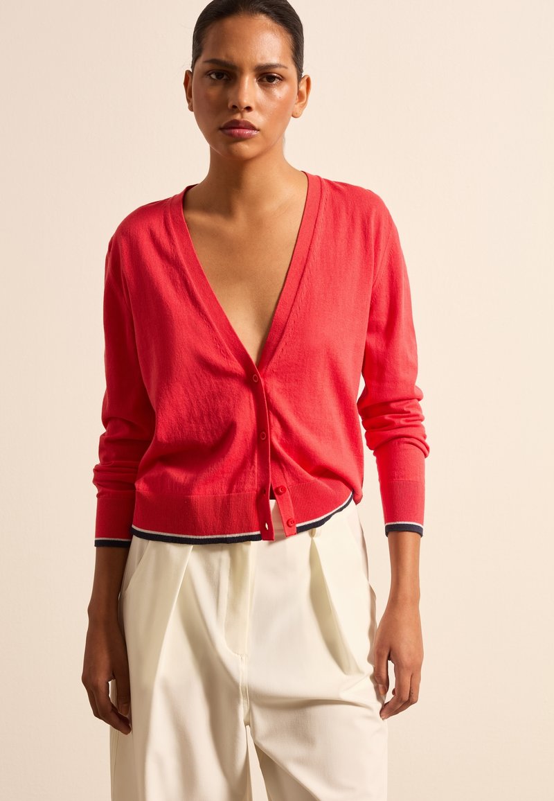 Roter Cardigan mit tiefem V-Ausschnitt, Knopfverschluss und navy gestreiften Bündchen. Getragen über crème-farbenen, plissierten Hosen. Weiche Textur.