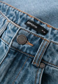 Jeans de mezclilla azul claro con detalles de costura en color cobre, cierre con botón metálico y etiqueta negra con el nombre de la marca "ombre."