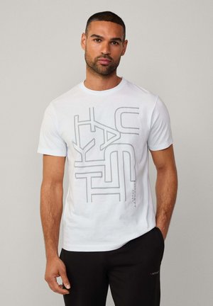 Weißes Baumwoll-T-Shirt mit einem grauen geometrischen Grafikdesign, kurzen Ärmeln und Rundhalsausschnitt. Kombiniert mit schwarzen Hosen.