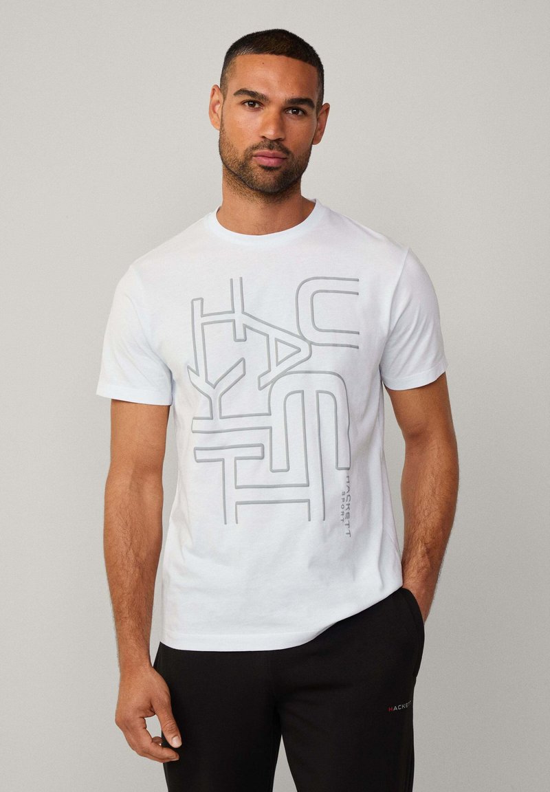 Witte katoenen T-shirt met een grijs geometrisch grafisch ontwerp, korte mouwen en een ronde hals. Gecombineerd met zwarte broek.