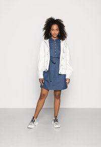 Casaco oversized branco sobre um vestido de camisa em denim azul com botões e cinto na cintura. Combinado com ténis brancos com detalhes em preto.