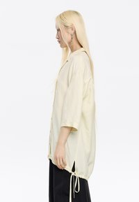Camicia oversize a bottoni gialla chiara, con maniche corte, orlo con coulisse e texture liscia. Design casual con una vestibilità rilassata.