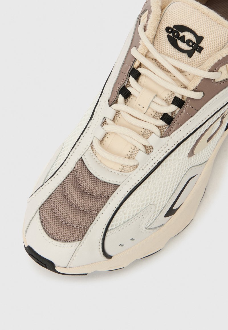 White and beige sneaker with mesh and leather materials. Features a rounded toe, textured accents, and a black logo on the tongue.