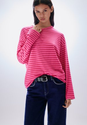 Pink striped long-sleeve top with a relaxed fit, paired with dark blue jeans featuring studded accents and a black belt.
