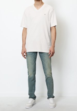 Dorosła osoba ubrana w białą koszulkę z krótkim rękawem i dekoltem w serek, wyblakłe niebieskie dżinsy oraz białe trampki, stojąca na tle jednolitej białej ściany.