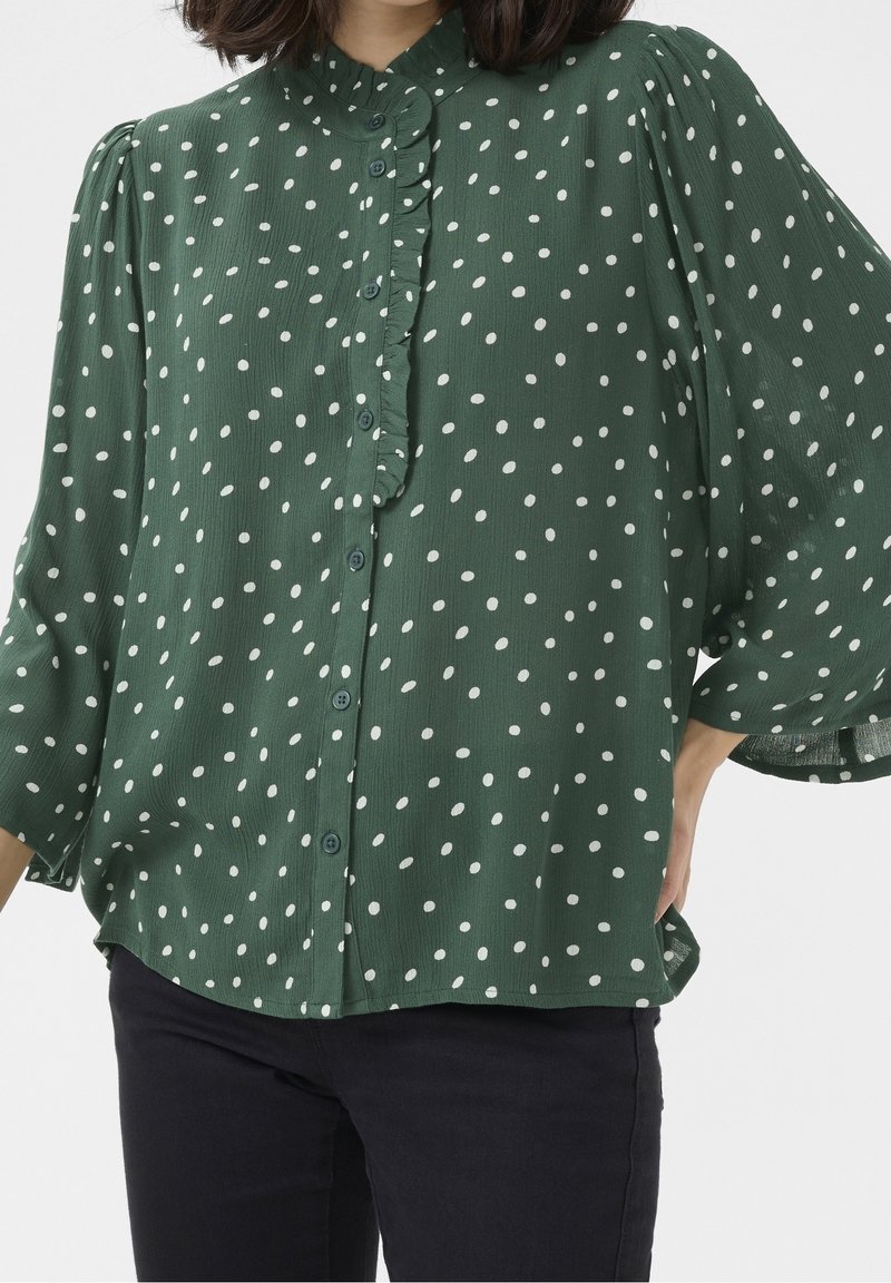Donna che indossa una blusa verde con pois bianchi, con una placca dei bottoni arricciata e maniche a sbuffo a tre quarti, abbinata a pantaloni scuri.