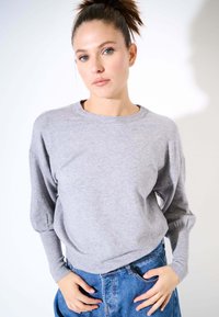 Felpa grigia a maniche lunghe con scollatura arrotondata e polsini elasticizzati, abbinata a pantaloni corti in denim a vita alta, su uno sfondo chiaro.