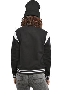 Svart sweatshirt med vita axelaccent, ribbade ärmar och nederkant samt en texturerad tyg. Bärs med en svart basker; grå jeans synliga.