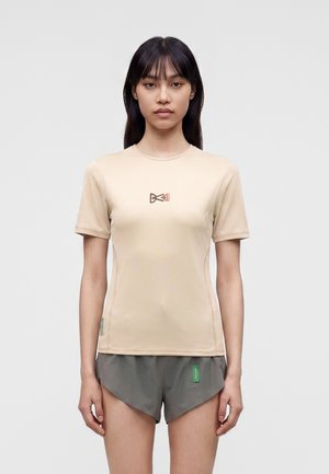 Ung kvinde med langt mørkt hår iført en beige kortærmet top med lille logo på brystet og grå træningsshorts, stående mod en ensfarvet baggrund.