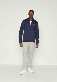 Sweatshirt zippé navy avec un col haut, fabriqué en tissu doux, présente un logo blanc sur la poitrine gauche. Assorti à un pantalon gris clair et des sneakers blanches.