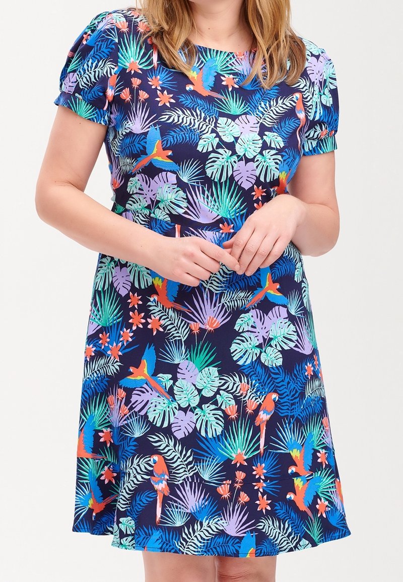 Frau in einem navyblauen Kleid mit bunten tropischen Blättern und Papageien, kurzen Puffärmel und Rundhalsausschnitt.