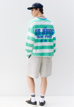 Jeune homme portant un t-shirt rayé vert et blanc avec un texte bleu en français, un short gris clair, une casquette noire, des chaussettes blanches et des baskets noires, debout de côté.