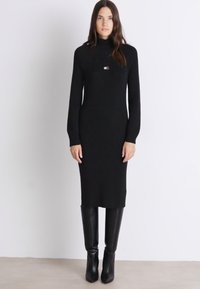 Svart ribbad stickad klänning med hög krage, slim fit och långa ärmar, med en liten logotyp på bröstet, tillsammans med figursydda svarta stövlar.