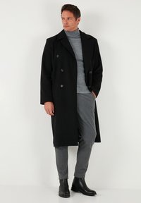 Cappotto doppiopetto nero in tessuto strutturato con grandi bottoni, indossato sopra un dolcevita grigio a coste e pantaloni grigi, abbinato a stivali neri.