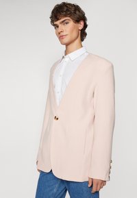 Hellrosa Blazer mit V-Ausschnitt, goldener Knopfverschluss und strukturierten Schultern. Kombiniert mit einem weißen Hemd und blauen Jeans.