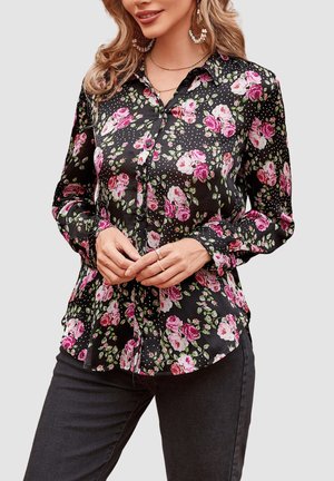Mujer con blusa negra abotonada con estampado floral en rosa y blanco y pantalones negros, manos entrelazadas al frente.