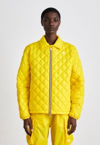 Casaco amarelo acolchoado com fecho frontal, padrão de losangos e pequeno logotipo branco. O tecido tem uma textura suave, complementado por calças coordenadas.