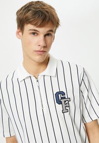 Valkoinen polo-paita, jossa on tummansinisiä pystysuoria raitoja, lyhyet hihat ja vetoketjullinen kaulus. Varustettu sinivihreällä brodeeratulla "CF"-logolla.