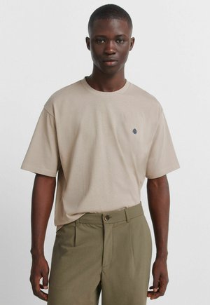 Jonge man draagt een beige T-shirt met korte mouwen en een klein donker logo op de linkerborst, samen met olijfgroene broek, staand tegen een effen lichte achtergrond.