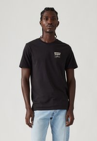 Juoda medvilninė marškinėliai su apvalia iškirpte, turintys mažą baltą "Levi's California" logotipą kairiajame krūtinės šone. Dėvimi kartu su šviesiai mėlynomis džinsais.
