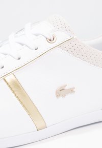 Bílá teniska se zlatými akcenty, s perforovanou béžovou patou, plochými tkaničkami a texturovaným povrchem. Logo viditelné na boku.