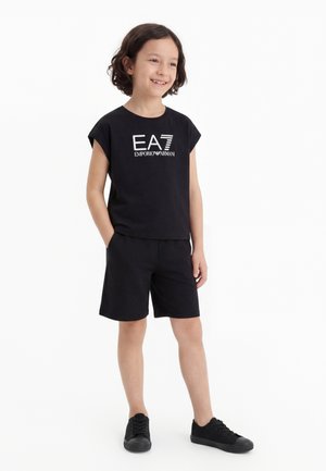 Copil zâmbitor cu păr scurt și închis la culoare, purtând un tricou negru EA7 Emporio Armani, pantaloni scurți negri și adidași negri, stând cu mâinile în buzunare.