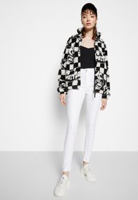 Camisola de fleece em padrão xadrez preto e branco com logótipos, combinada com jeans skinny brancos e sapatilhas claras. Um top preto e um boné completam o visual.