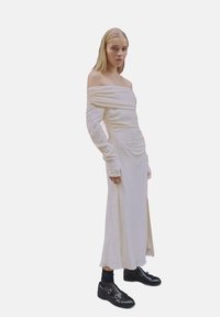 Off-shoulder vit klänning med en rynkad design, långa ärmar och golvlång nederdel. Buren med svarta skor med dubbla spännen.
