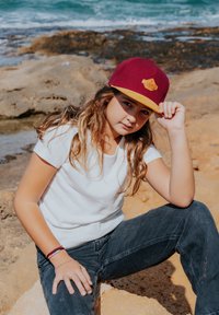 Bordeaux hat med en gul skygge og logo, båret med en hvid t-shirt og sorte jeans. Baggrunden har en stenstrand og hav.