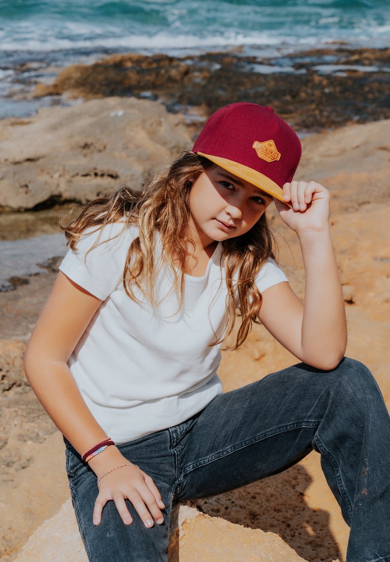 Bordeaux hat med en gul skygge og logo, båret med en hvid t-shirt og sorte jeans. Baggrunden har en stenstrand og hav.
