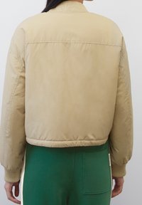 Giacca bomber beige con colletto a coste, texture imbottita e lunghezza accorciata. Le maniche presentano una leggera arricciatura ai polsini.