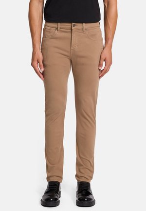 Pantaloni slim-fit color marrone chiaro, realizzati in cotone, con chiusura a zip e bottone metallico, cinque tasche e dettagli di cucitura puliti. Indossati con scarpe nere.