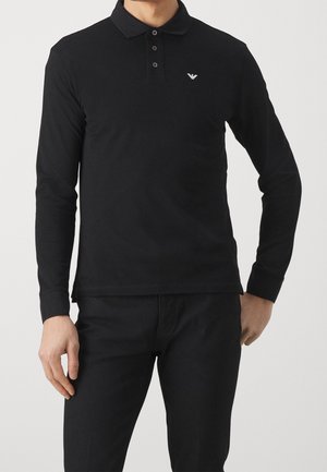 Mann trägt ein schwarzes Polohemd mit langen Ärmeln, drei Knöpfen und einem kleinen weißen Logo auf der Brust, kombiniert mit schwarzen Hosen.