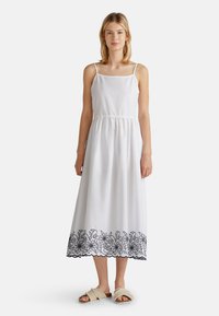 Weißes Baumwollkleid mit Spaghettiträgern, gekräuseltem Taillenbereich und schwarzer Blumenstickerei am Saum. Kombiniert mit beigen Rutschsandalen.