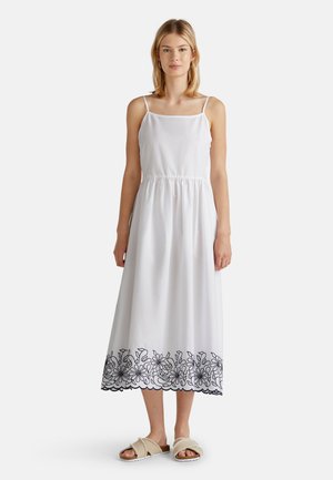 Vestido de algodón blanco con tirantes finos, cintura fruncida y bordado floral negro en el dobladillo. Combinado con sandalias deslizantes beige.