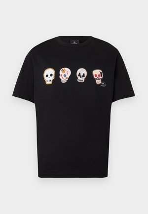 Jeune homme debout les mains dans les poches, portant un t-shirt noir avec quatre crânes colorés, un pantalon beige et des baskets blanches.