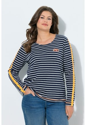 Kvinde med langt bølget hår iført navy- og hvidstribet langærmet skjorte med gule kringlemønstrede striber på ærmerne og blå jeans.