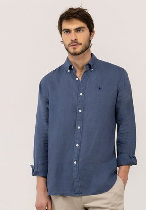 Hombre con pelo castaño, viste una camisa azul de manga remangada y pantalones beige sobre un fondo liso.