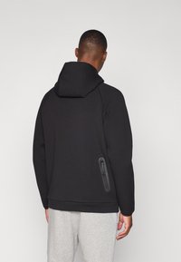 Sweatshirt à capuche noire en tissu doux, avec une poche zippée sur le côté gauche et des poignets côtelés, associé à un pantalon gris clair.