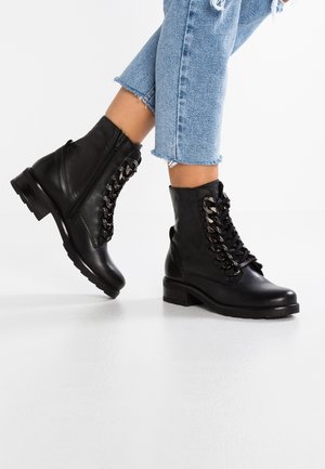 Czarne skórzane botki za kostkę z masywnym łańcuchem, noszone z jasnoniebieskimi, krótkimi, postrzępionymi dżinsami na prostym białym tle.