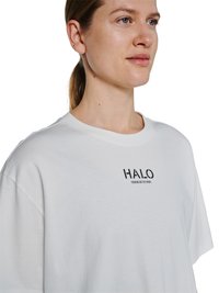 Junge Person mit heller Haut und zurückgebundenem Haar trägt ein weißes T-Shirt mit schwarzem "HALO"-Logo auf der Brust, schaut leicht nach rechts vor weißem Hintergrund.