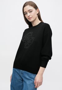 Melnā džemperī ar teksturētu logotipa dizainu priekšpusē. Piegriezums ir vaļīgs ar nolaistām plecu līnijām un ribotām aprocēm, kombinēts ar zilām džinsa biksēm.