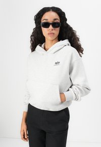 Žena s dlhými kučeravými vlasmi, nosiaca čierne slnečné okuliare, svetlošedú mikinu Alpha Industries a čierne nohavice, stojaca na bielom pozadí.