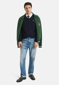 Grüne Jacke mit weißen Streifen, marineblauer Pullover, weißes Hemd, legere blaue Jeans, schwarze Schuhe und ein Logo auf der Jacke.